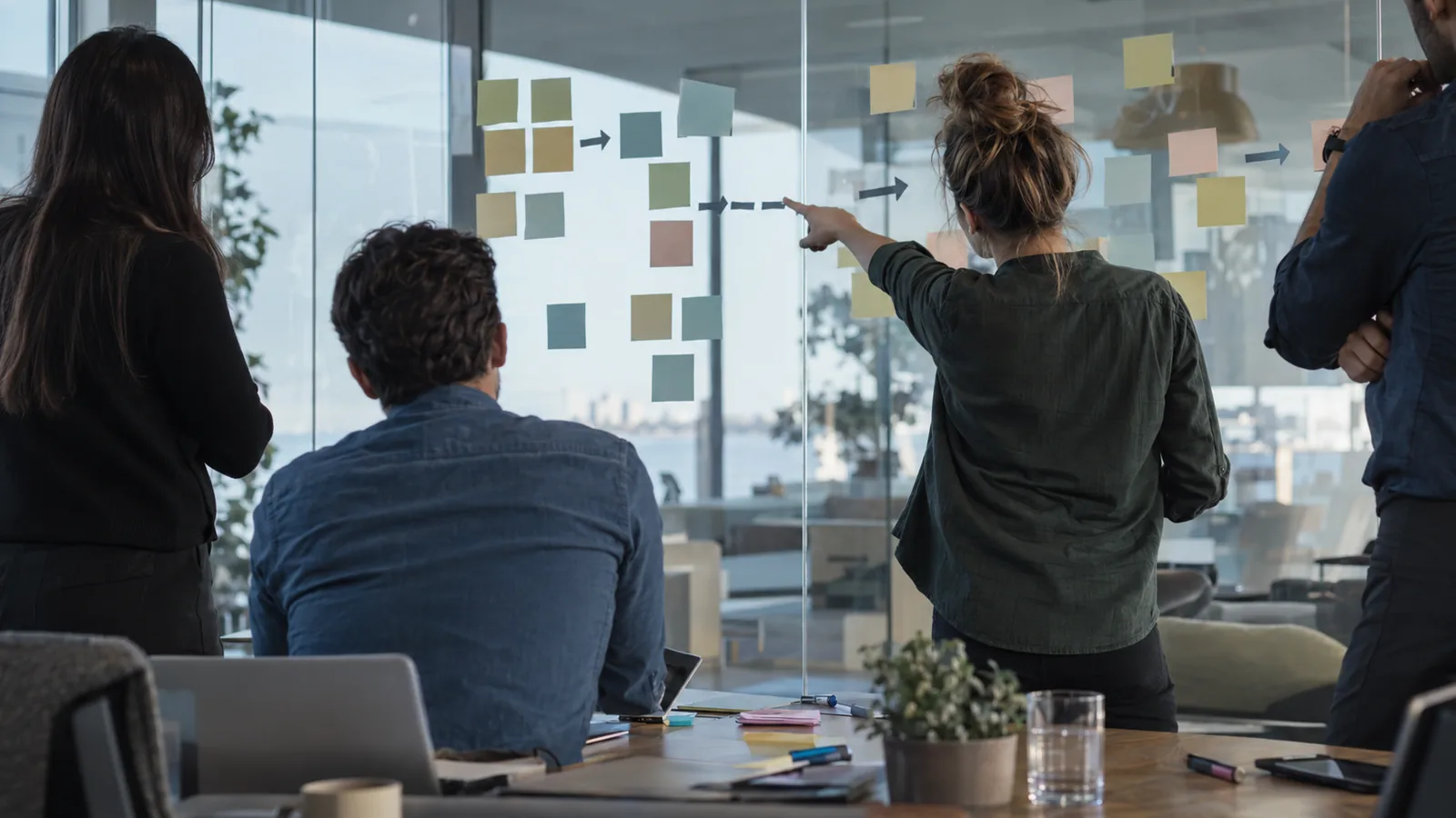 A modern meeting room scene shows a consultant and team members viewed from behind mapping a workflow on a glass wall covered with blank sticky notes, tape arrows, marker smudges, and scattered uncapp